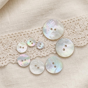 May include: A collection of iridescent, round mother-of-pearl buttons in various sizes. Each button has two holes for sewing. The buttons are arranged on a beige fabric with a delicate lace trim, creating a soft, natural aesthetic.