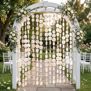 Peut inclure: Une arche de mariage en bois blanc ornée de roses blanches et de feuillage vert, avec des brins suspendus de disques circulaires iridescents ressemblant à des coquillages. L'arche se dresse sur un chemin en pierre dans un jardin.