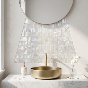 May include: A round gold vessel sink sits on a white marble countertop in front of a backsplash made of iridescent white mother-of-pearl mosaic tiles. A round mirror is mounted above the tiles, and a white soap dispenser is to the left.