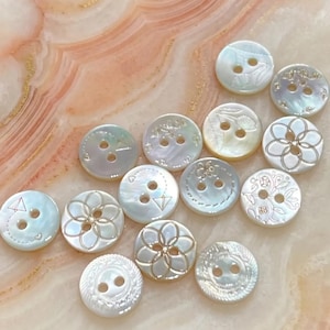 Puede incluir: Colección de botones iridiscentes de nácar con diversos diseños florales y geométricos grabados. Cada botón tiene dos o cuatro agujeros para coser. Los botones son en tonos de blanco, crema y azul pálido.