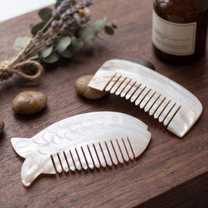 Puede incluir: Dos peines para el cabello hechos de material de concha blanca iridiscente. Un peine presenta un borde curvo y liso, mientras que el otro tiene forma de pez con detalles de escamas talladas. Ambos peines tienen dientes finos y uniformes.
