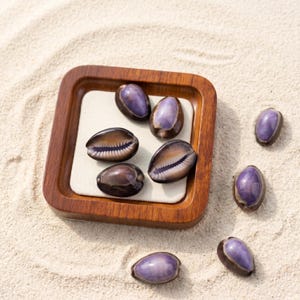 May include: A collection of polished, purple-hued cowrie shells, some displayed on a wooden tray with a white insert, resting on a bed of sand. The shells vary in size and have a glossy finish, showcasing natural patterns.