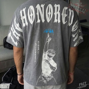 Puede incluir: Camiseta gris con la palabra "HONORED" en letras blancas grandes en la espalda. La camiseta presenta una ilustración blanca de una persona que señala hacia arriba, con texto japonés y detalles en azul. Las mangas tienen diseños blancos.