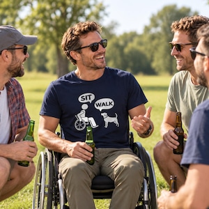 May include: A navy blue short-sleeved t-shirt featuring a white graphic print of a person in a wheelchair with a speech bubble saying SIT and a dog with a speech bubble saying WALK. The shirt is made of cotton and has a crew neckline.