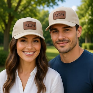 Op de afbeelding: Twee mensen dragen beige baseballpetten met een bruin leren patch. De patch heeft de Griekse letters "AΩ>i". De petten hebben een gebogen klep en een gestructureerde kroon. De achtergrond is een vage groene park.