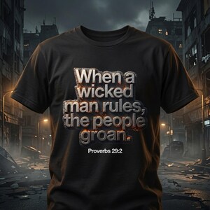 May include: A black short-sleeved t-shirt featuring the text "When a wicked man rules, the people groan." in a metallic, stone-like font with glowing orange edges. Below the main text, the citation "Proverbs 29:2" is printed in white.