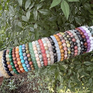 May include: A selection of beaded bracelets in diverse colours, such as green, blue, pink, and black. The bracelets are presented on a white cylindrical form, set against a backdrop of green foliage. The beads appear to be made of stone.