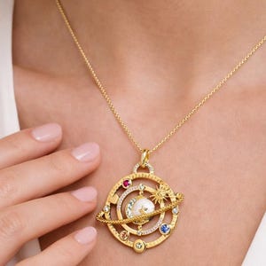 May include: Gold-toned celestial pendant necklace. The pendant features a planet design with colorful gemstones and a central pearl. The necklace is worn on a delicate gold chain.