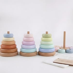 Op de afbeelding: Drie houten stapelspeelgoed met pastelkleurige ringen en een houten pin. Het speelgoed is in tinten oranje, roze en groen. Een witte stoffen zak en een houten label met tekst staan op de voorgrond.