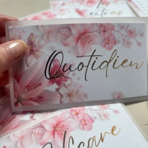 Könnte beinhalten: Eine rechteckige Karte mit einer halbtransparenten, mattierten Auflage, die ein rosa Blumenmuster aus Lilien und Kirschblüten zeigt. Das Wort Quotidien ist in einer schwarzen Schreibschrift in der Mitte der Karte über die Blumen gedruckt.