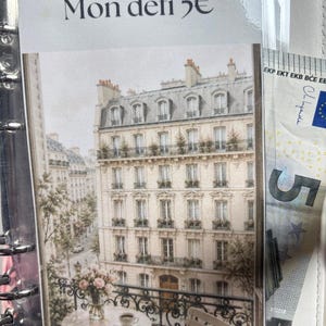 Könnte beinhalten: Eine laminierte Karte mit dem Text Mon défi 5€ und einer Illustration eines Pariser Gebäudes mit Balkon, einem kleinen runden Tisch, einer Blumenvase und einer Tasse. Ein Fünf-Euro-Schein steckt seitlich in der Ringbuchmechanik.