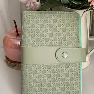 Peut inclure: Un agenda vert sauge avec un motif tissé et une fermeture à sangle assortie. L'agenda a un bord vert menthe et une fermeture à bouton. Il est devant un pichet blanc avec des fleurs roses.
