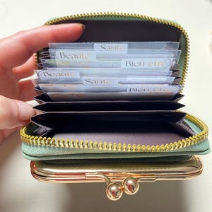 以下が含まれることがあります： ゴールドのジッパーと留め金が付いた、開いたセージグリーンの財布。財布には、「Beaute」、「Sante」、「Bien être」とラベル付けされたカードディバイダーが詰められています。