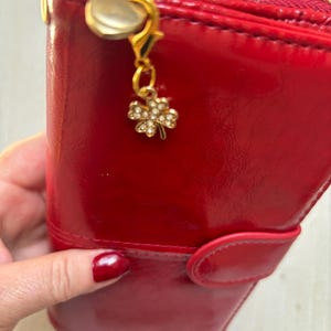 May include: A glossy red wallet featuring a gold-toned zipper closure and a small, dangling charm shaped like a four-leaf clover. The clover charm is embellished with tiny, clear rhinestones, and the wallet includes a snap-button flap closure.