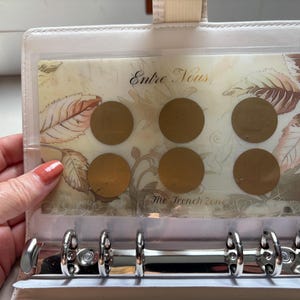 May include: A white six-ring binder insert featuring a translucent sheet with a botanical leaf pattern in shades of brown and cream. The sheet displays the text "Entre Nous" and "The Trench Zone" alongside six circular gold-coloured foil stickers.