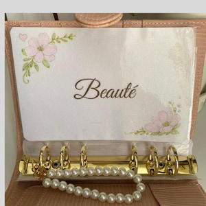 Peut inclure: Un agenda rose avec des anneaux dorés et un bracelet de perles. L'agenda contient une carte avec le mot "Beauté" et des motifs floraux. Le texte "THE FRENCHZONE" est en bas.
