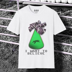 May include: A white short-sleeved t-shirt featuring a graphic of a bright green cone shape topped with a dark patterned bow. Inside the cone is a small black silhouette of a creature. The text I WANT TO BELIEVE is printed in black below the graphic.