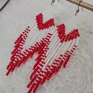Könnte beinhalten: Weiß-rote Perlenohrringe mit Fransenmuster. Die Ohrringe haben ein herzförmiges Muster an der Oberseite und eine Kaskade aus roten Perlen. Sie sind an silberfarbenen Ohrhaken befestigt.