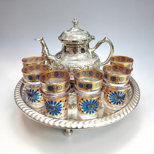 Puede incluir: Un juego de té plateado con una tetera y ocho vasos, todo sobre una bandeja plateada. Los vasos tienen diseños florales azules y dorados. La tetera y la bandeja tienen detalles ornamentados.