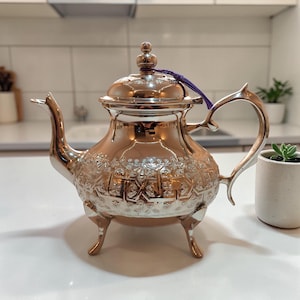 Puede incluir: Una tetera plateada con intrincados diseños florales y geométricos. La tetera tiene un pico curvo, un asa decorativa y una tapa con un pequeño pomo. Una cuerda morada está unida a la tapa. La tetera se apoya sobre tres patas pequeñas.