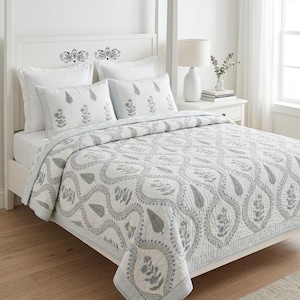 Könnte beinhalten: Ein weiß-blaues Bettwäsche-Set mit floralem Muster auf einem Bett. Die Steppdecke zeigt ein sich wiederholendes Design aus stilisierten Blättern und Blumen in Blau- und Grautönen. Das Set enthält passende Kissenbezüge und eine Tagesdecke.