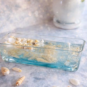Peut inclure: Un bougeoir rectangulaire en verre rempli de cire bleue et transparente, de coquillages et de paillettes dorées. Deux mèches blanches sont visibles. Plusieurs coquillages sont dispersés.