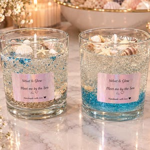 May include: Two clear glass candles filled with gel wax, blue and silver glitter, and small seashells. Each candle features a rectangular iridescent label with the text Velvet & Glow, Meet me by the Sea, and Handmade with love.