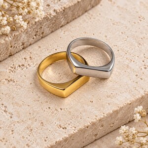 Peut inclure: Deux bagues chevalières, l'une en ton or poli et l'autre en ton argent poli, présentant une surface supérieure rectangulaire lisse et plate ainsi qu'un anneau arrondi. Les bagues sont placées ensemble, celle en argent reposant sur celle en or.