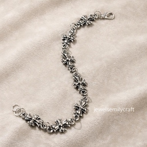 May include: A silver bracelet with a series of decorative cross-shaped links. The bracelet has a lobster clasp and a small loop. The jewellery is displayed on a neutral, textured surface.