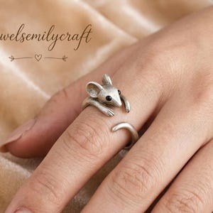 May include: A silver-colored mouse ring with black eyes and a curved tail. The mouse wraps around the finger, with its paws resting on the finger. The ring is worn on a hand.