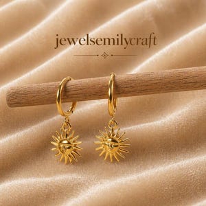 Puede incluir: Pendientes de aro dorados con colgantes de sol. Los pendientes cuelgan de una clavija de madera sobre un fondo beige suave. Los colgantes de sol tienen una cara y rayos puntiagudos. El texto "jewelsemilycraft" está encima.