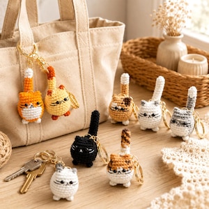 May include: A collection of small, round, crocheted cat keychains in various colours including orange, yellow, black, white, grey, and calico. Each cat features a long, upright tail, embroidered facial details, and a gold-toned metal keyring.