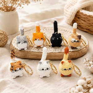 May include: A collection of eight small, round, crocheted cat keychains in various colors including gray, orange, black, calico, white, and yellow. Each cat features a tall, upright tail, embroidered facial details, and a gold-toned metal keyring.