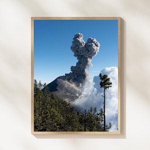May include: A framed print of a volcanic eruption. The image shows a mountain with a dark, rocky slope, topped by a large plume of gray smoke and ash against a blue sky. Green trees are in the foreground.