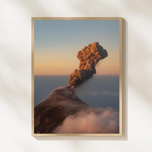 May include: A framed print of a volcanic eruption. The image captures the volcano's peak with a plume of smoke and ash against a sunset sky. The artwork is encased in a wooden frame, adding a natural aesthetic to the landscape photograph.