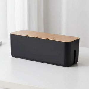 May include: A black cable tidy box with a light brown bamboo lid. The rectangular box has three small openings on the top and a slot on the side for cable organisation. The box is on a white surface.