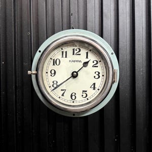 May include: A round, light blue wall clock with a silver-colored bezel and a white face. The clock has black numerals and hands, with the brand name "KAPPA" printed at the top. The clock is mounted on a black, vertically-grooved surface.