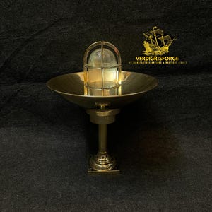 May include: A brass nautical-style light fixture with a round, bowl-shaped shade and a caged glass enclosure. The fixture is mounted on a pedestal base. The background is a dark, textured surface.
