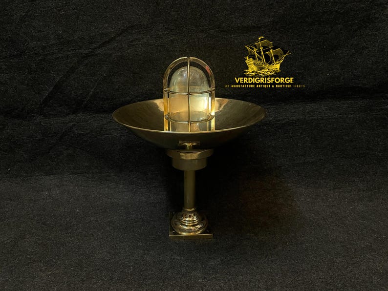 May include: A brass nautical-style light fixture with a round, bowl-shaped shade and a caged glass enclosure. The fixture is mounted on a pedestal base. The background is a dark, textured surface.