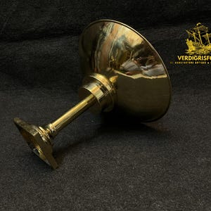 May include: A polished brass pedestal bowl with a wide, shallow bowl and a slender stem. The bowl is angled, reflecting the surrounding environment. The base is square. The logo "VERDIGRISFORGE" is in the upper right corner.