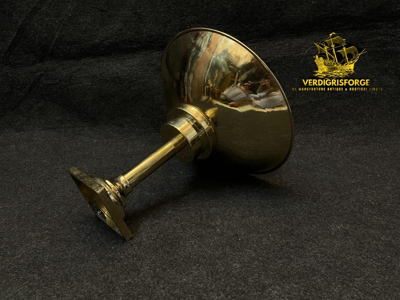 May include: A polished brass pedestal bowl with a wide, shallow bowl and a slender stem. The bowl is angled, reflecting the surrounding environment. The base is square. The logo "VERDIGRISFORGE" is in the upper right corner.