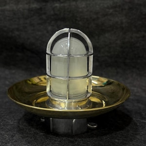 Puede incluir: Una lámpara de latón y vidrio de estilo vintage. La lámpara presenta una cúpula de vidrio transparente encerrada en una jaula de metal, montada sobre una base circular de latón. La base tiene un diámetro de aproximadamente 20 cm. La luz está diseñada para uso en interiores o exteriores.