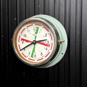 May include: A round clock with a white face, red and green accents, and black hands. The clock has a silver bezel and a light green backing. The clock is mounted on a black, vertically-lined surface.