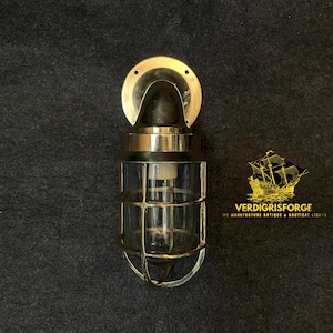 Op de afbeelding: Een nautische wandlamp van messing en glas met een beschermende kooi. De lamp heeft een ronde montageplaat en een heldere glazen behuizing. De afbeelding bevat ook de tekst "VERDIGRISFORGE" en een scheepslogo.
