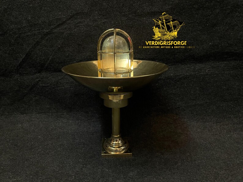 May include: A brass nautical-style wall sconce with a caged glass light fixture. The sconce has a round, bowl-shaped reflector and a decorative base. The text "VERDIGRISFORGE" and a ship logo are in the upper right corner.