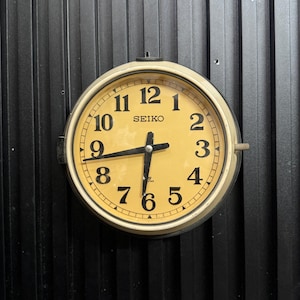 May include: A round, vintage Seiko wall clock with a cream-colored face and black numerals. The clock has black hands and a metal frame. The clock is mounted on a black, vertically-lined surface.