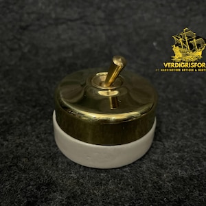 Op de afbeelding: Een messing en witte tuimelschakelaar. De schakelaar heeft een ronde messing bovenkant met een messing tuimelaar en een witte basis. Het logo "VERDIGRISFORGE" is zichtbaar in de rechterbovenhoek.