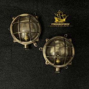 May include: Two round brass nautical bulkhead lights featuring a protective metal cage over a clear glass dome. Each fixture has a brass base with mounting tabs and a central bulb socket. The logo for VERDIGRISFORGE with a ship icon is visible.