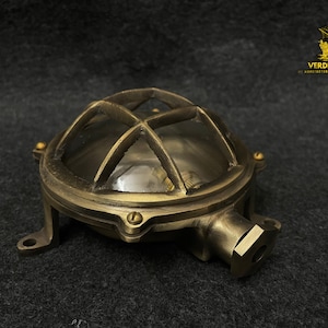 May include: A round brass bulkhead light fixture featuring a protective metal cage over a clear glass dome. The fixture has a weathered metallic finish, mounting tabs with screw holes, and a side-mounted conduit entry point for electrical wiring.