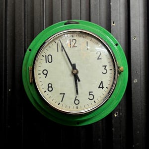 Può includere: Un orologio rotondo con quadrante bianco e numeri neri, racchiuso in una cornice verde brillante. Le lancette dell'orologio indicano circa le 5:56. L'orologio è montato su una superficie nera strutturata.